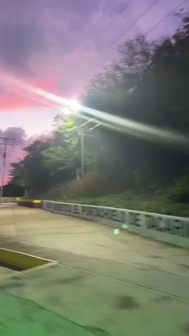 The video captures a moving shot of a road at sunset. The sky is a vibrant blend of purple and pink hues with dramatic clouds. Power lines and streetlights are visible, as is the silhouette of trees and hills in the background. In the foreground, a road surface with painted markings and bollards is present. A sign with the word GOMEZ is also visible. The scene appears to be a scenic route or tourist area, with the Venezuelan flag colors incorporated into some of the markings. The motion suggests the video was taken from a vehicle.