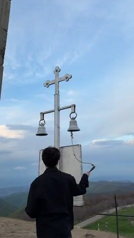 The video shows a person ringing church bells. The individual, wearing a black jacket, stands facing a metal structure. The structure consists of a cross at the top, two hanging bells, and a white sign below. A gray concrete pillar stands to the left of the metal structure. The person pulls a rope, ringing the bells. The camera pans up along the pillar, revealing a small shrine mounted on it. The camera then briefly focuses on another person with long dark hair, looking out at a scenic view of mountains and a sky with scattered clouds.