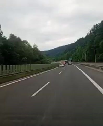 The video shows a first-person perspective of driving on a highway. The road is asphalt with white lines marking the lanes. On the left side of the road, there is a metal guardrail and trees. On the right side, there are more trees and a highway sign that says "Zagorje" with a distance of 7 km. Several cars are visible in front of the vehicle, and the sky is cloudy. The overall environment appears to be a wooded, hilly area.