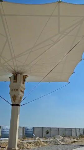 The video displays a structure resembling the Kaaba, a cube-shaped building central to Islam, under a bright blue sky with sparse white clouds. The structure is primarily black with gold decorative bands and inscriptions. White fabric awnings, supported by metal frames, are visible in the foreground and background, providing shade. The scene appears to be an outdoor area, possibly a courtyard or plaza, with a low concrete wall and some vegetation in the distance. The sun is shining brightly, casting shadows. The camera pans slowly across the scene. No people are visible in the video.