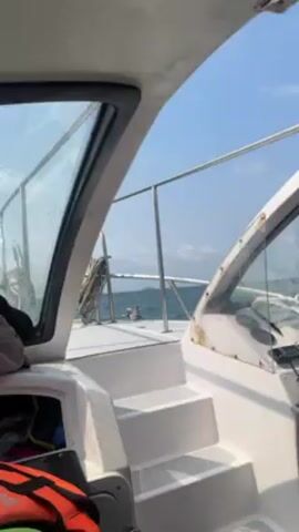 The video shows the view from inside a boat, looking out over the water. The boat appears to be moving, with water visible through the windows. A metal railing is visible on the side of the boat.  Inside, there are white steps leading to a different level, and what appears to be a life jacket is visible in a compartment. The sky is blue and sunny. A small boat can be seen in the distance.