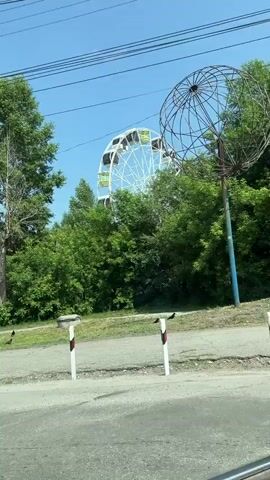 The video shows a view from a moving vehicle, looking towards a Ferris wheel partially obscured by trees. The Ferris wheel is white with multiple cabins. There are also trees, power lines, and a clear blue sky visible. In the foreground, there are three white posts with red and white reflective markers, and a paved road. There are also some dark birds on the grass. The text overlay identifies the location as Gorky Park, Leninsk-Kuznetsky.