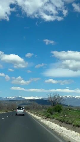 The video shows a view of a road with a silver car driving on it, mountains in the background covered with snow, and a bright blue sky with fluffy white clouds.  A bare tree is visible along the roadside. The camera appears to be positioned inside a car moving along the road. The view is mostly sky and landscape.