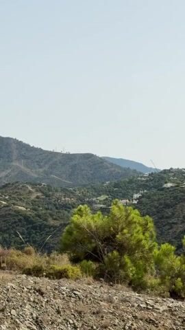 A slow panning shot moves across a lush, hilly landscape under a bright, clear sky. In the foreground, a rocky, dry patch of ground is dotted with sparse vegetation, including a vibrant green pine tree. The middle ground is dominated by rolling hills covered in dense green foliage, interspersed with scattered white buildings, suggesting a populated area or settlement. Some of these buildings appear to be on the slopes of the hills. Further in the distance, more hills recede into the background, with faint structures visible on their crests. The overall impression is of a sunny day in a scenic, possibly Mediterranean, rural or semi-rural environment. There are no people or overt actions depicted in the video.