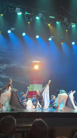 A group of approximately twelve dancers performs on a dimly lit stage featuring a large red and white striped lighthouse prop in the center. The stage is illuminated by overhead blue and yellow spotlights, and the backdrop displays a textured image of dark blue ocean waves. The dancers, predominantly female, wear black tops and flowing skirts or wide-leg pants in black, white, or light blue. They execute contemporary dance movements, including kneeling, bending forward, and sweeping arm gestures, with some dancers moving from a kneeling to a standing or partially standing position. The lighthouse prop emits a warm yellow glow from its top section. The heads of a few audience members are visible in the lower foreground.