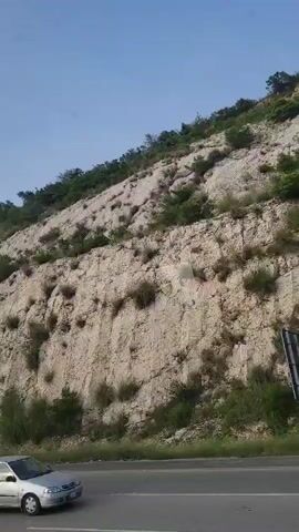 The video shows a road running along the base of a rocky, vegetated hillside under a clear blue sky. The hillside is steep and appears to be a natural rock face with some areas of erosion and smaller rocks. Green vegetation, including bushes and grasses, grows in patches on the hillside and at its base. A paved road with white lane markings is visible in the foreground. The perspective suggests the video was taken from a moving vehicle.