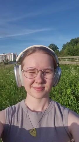 A young woman wearing glasses and large silver headphones is outside on a sunny day. She is smiling and winking at the camera. The background is a grassy field with trees and a building in the distance. The video is a selfie, as the camera appears to be held by the woman.