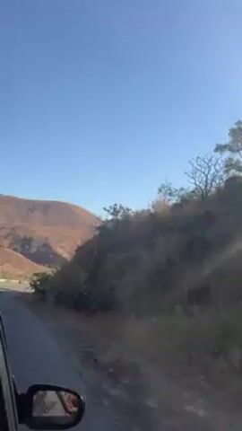 The video shows a view from inside a moving vehicle, looking out at a winding road that leads into a hilly landscape. The hills are brown and sparsely vegetated, suggesting a dry climate. The sky is clear and blue. The car's side mirror is visible in the foreground, and the road ahead has some white and green markings along the edges. The camera appears to be held still as the car moves along the road.