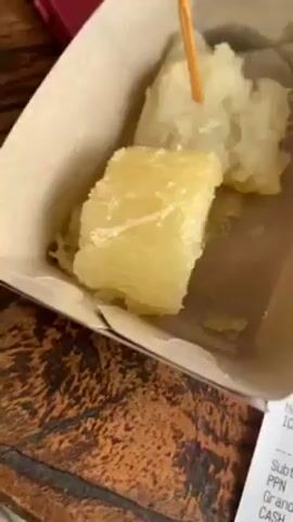 Here is a detailed description of the video:

The video presents a still shot of a meal consisting of fried cassava and iced hazelnut coffee, alongside a receipt from Kedai Bukit Rhema. The cassava, served in a small cardboard container and held together by a toothpick, shows a slightly oily sheen. Next to it is a plastic cup of iced hazelnut coffee, featuring a black straw. Droplets of condensation cling to the exterior of the cup.

The receipt details the purchase: "Hazelnut Coffee ICE" costing Rp 30,000. Adding 10% PPN (tax) brings the grand total to Rp 33,000. The customer paid with Rp 35,000, receiving Rp 2,000 in change. The receipt also includes the business name, “Kedai Bukit Rhema,” their contact information for booking sunrise and meals, the date and time of the sale (May 11, 2023, at 09:39), the cashier’s name (Devy), and a thank you note expressing gratitude for supporting UMKM Borobudur Magelang.

The meal and receipt are positioned on a wooden table with a textured, slightly worn surface, adding to the rustic feel of the scene. A red phone is seen in the background of the image. The overall lighting emphasizes the colors of the items on the table.