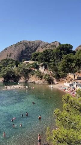 The video shows a body of water, likely a bay or cove, with several people swimming and wading in the clear turquoise water. The water is shallow near the shore, revealing the rocky seabed. A small beach area with sunbathers is visible on the right side of the frame, backed by a cluster of buildings and lush green trees. In the background, a large, rocky mountain with sparse vegetation rises dramatically against a clear blue sky. The overall scene depicts a sunny day at a popular coastal recreation area.