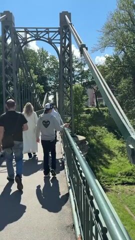 Four people are walking across a green metal bridge on a sunny day. The bridge has a lattice structure and is bordered by a railing. Trees and foliage are visible on either side of the bridge, and a building can be seen in the distance. The people are walking away from the camera and appear to be in casual clothing. The person in front is wearing a white coat, the person next to them is wearing a grey hoodie with a heart design and a white baseball cap, and the two people further back are wearing dark clothing. Shadows are cast on the bridge surface by the people and the bridge structure.