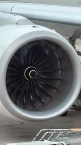 The video shows a static close-up view of the engine of a white airplane. The engine has a silver casing and black turbine blades visible inside. A white spiral design is visible at the center of the turbine. A portion of the airplane's wing is visible above the engine. In the lower part of the frame, part of a white cart with a ladder is visible. The overall environment appears to be an airport tarmac. There is some slight movement or heat haze visible inside the engine.