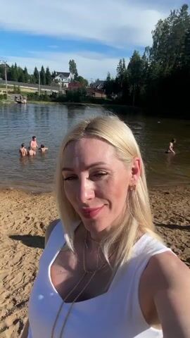 The video shows a medium shot of a blonde woman with bright pink lipstick and a white V-neck top, wearing layered gold necklaces. She is standing on a sandy beach, with a lake behind her. The lake has a few people swimming in it and a red and yellow inflatable raft near the shore. A line of trees is visible across the lake, and beyond that, houses and a few power lines. The sky is blue with scattered white clouds. The woman is looking at the camera and appears to be speaking, but there is no audio. The camera angle is slightly upward, giving a close-up view of her face. The video is short, lasting only a few seconds, and shows minimal movement.