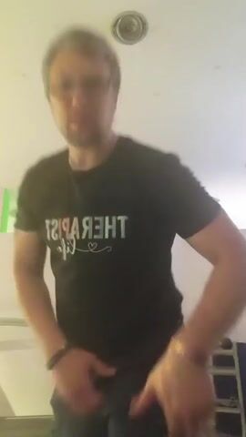 The video shows a person in a black t-shirt and black pants doing high knees in a gym or exercise room. The person is wearing glasses and has a watch on their left wrist. In the background, there is a treadmill, some weights on a rack, and ceiling lights. The person moves their arms and legs in a rhythmic motion as they perform the exercise. The video captures the movement from the chest up, with the person's face blurred due to motion.
