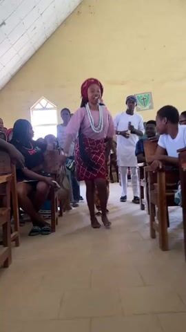 A young woman wearing a pink top, a red headwrap, a patterned red skirt, and beaded necklaces is dancing in what appears to be a church or community hall. She is barefoot and holding a whisk or fly swatter. Her movements are energetic and joyful. She is surrounded by other people sitting on wooden benches, watching her perform. Some of the spectators are smiling and appear engaged in the performance. The room has a sloped ceiling with exposed beams and fluorescent lighting. A cross emblem is visible on the wall in the background. The scene is brightly lit, suggesting it is daytime. The overall atmosphere seems festive and celebratory. The location is Dutsen Kato, Nigeria.