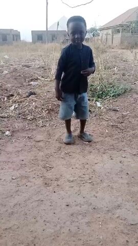 A young child stands in a dirt yard wearing a dark, long-sleeved shirt, denim shorts, and gray shoes. Buildings are visible in the background, and there is scattered debris on the ground around the child. The child looks directly at the camera.
