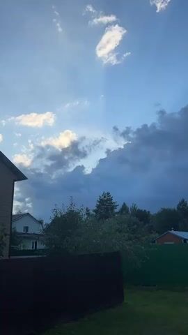 A video shows a partially overcast sky with dramatic clouds. Sunlight is breaking through the clouds in some areas, creating bright rays. Below the sky are trees and the tops of houses and fences. One house is light green and white with windows. Another house has a reddish-brown roof and blue siding. There is a dark brown fence in the foreground.