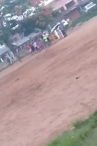The video shows a dirt road next to a green grassy area. There are people and vehicles on the left side of the road. The camera is unstable and moves around.