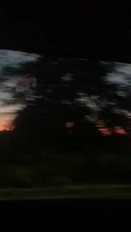 The video shows a sunset view from a moving vehicle. The sky transitions from a dark blue to a deep orange and red hue near the horizon. Silhouettes of trees and power lines are visible against the twilight sky. The motion blur suggests the camera is in motion, likely looking out a window. The overall scene is dark, with the vibrant colors of the sunset being the main focal point.
