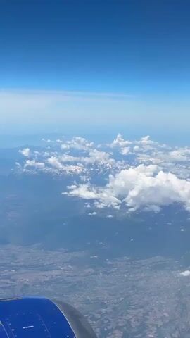 The video captures an aerial view from an airplane window. The sky is a clear, bright blue with some wispy clouds near the horizon. Below the clouds, a mountain range is visible, with patches of snow on the peaks. The mountains are partially obscured by fluffy white clouds scattered across the landscape. Further below, a vast expanse of land is seen, featuring a patchwork of fields, scattered buildings, and what appears to be a town or city. In the foreground, the blue and gray engine of the airplane is partially visible, indicating the perspective of the viewer. The overall scene is one of a scenic flight over a mountainous region.