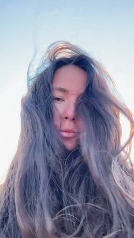 This is a close-up, low-angle video of a woman with long, dark, wavy hair. She is outdoors, possibly on a boat, as there is water and a shoreline visible in the background, along with a bright sky. The woman is looking at the camera and appears to be smiling slightly. The lighting suggests it is daytime, possibly late afternoon or early morning, with the sun creating a lens flare effect. Her hair is blowing in the wind, framing her face. She is wearing a white top. The video is short, lasting only a few seconds.