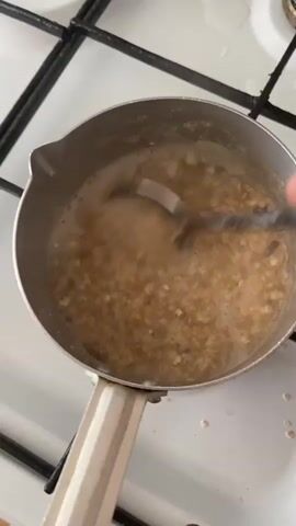A close-up shot shows a small metal pot with a handle sitting on a white gas stove. Inside the pot, a light brown, foamy liquid is being stirred with a spoon, creating a swirling motion. The liquid appears to be cooking oatmeal or a similar porridge. The stove has black grates and white enamel. The shot is slightly blurry due to the movement.