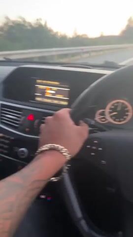 The video shows a person driving a Mercedes. The camera is positioned from the driver's perspective, focusing on the steering wheel and dashboard. The driver has a tattoo on their left arm and is wearing a gold bracelet. The speedometer shows the car is moving at around 74 mph. The environment outside the car is a highway with other cars visible. The sky is bright, suggesting it is daytime or early evening. Music is playing in the background.