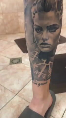 The video presents a detailed close-up of a person's lower leg, which is adorned with an intricate, full-calf tattoo in black and grey. The tattoo features several distinct elements: a portrait of a woman with an intense gaze, an older, bearded figure (possibly a mythical deity or ancient warrior), a Viking longship sailing on waves, a chalice or cup, and an eye with radiating lines at the bottom. Interspersed throughout are geometric and symbolic patterns, resembling runes or other ancient script. Near the ankle, the phrase "Suerte en grande" is clearly visible in elegant script, with "Suerte en gracia" also partially visible at another point. The tattooed skin appears slightly wet or recently moisturized, giving it a glossy sheen. The person is wearing a grey slip-on sandal. The background consists of a light-colored tiled floor with a grid pattern, and two dark floor mats are visible on the floor. The camera slowly pans around the leg, allowing for a comprehensive view of the detailed artwork.