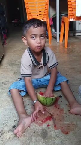 A young boy is sitting on the floor eating watermelon. He is wearing a tan polo shirt with a black collar and blue shorts. The watermelon is in a hollowed-out rind and has been mostly eaten, with juice and seeds spilled on the floor around him. A spoon is lying next to the watermelon. The boy is looking up and to his left, and appears to be talking or reacting to something off-camera. He is sitting on a concrete floor in what appears to be a home or covered outdoor area, with orange plastic chairs visible in the background.