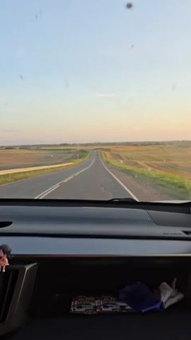 The video is shot from the interior of a car looking out the windshield. The car is driving on a paved road through a rural landscape. The road is a two-lane highway with white lines marking the lanes. On either side of the road are fields of dry grass and some scattered trees. The sky is clear and blue with a hint of orange from the setting sun. The sun is low on the horizon, casting long shadows. There are other cars on the road ahead. The dashboard of the car is visible at the bottom of the frame.