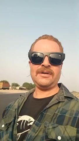 The video features a man standing near a highway. He wears a black t-shirt with a white graphic, a green and black plaid shirt, and dark sunglasses. He also has a mustache. Behind him, trucks are parked on the highway. The sky is clear, and the environment appears to be sunny. The camera seems to be handheld, recording the man, possibly in a selfie mode. The man is looking directly at the camera.