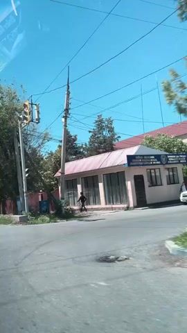 The video shows a street scene on a sunny day. A four-way traffic light hangs above an intersection, displaying a yellow light. A man is walking on the sidewalk near a low, long building with a pink roof and many windows. Power lines are visible overhead, connected to utility poles. A large tree stands on the side of the road. The video is taken from a moving vehicle. The road is asphalt.