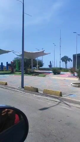 The video shows a view from inside a moving vehicle, likely a car, of a waterfront scene. The vehicle is traveling along a road with a painted median barrier in yellow. On the other side of the road is a grassy area with various features.

The grassy area includes several large white canopy structures that provide shade, as well as planted trees, bushes, and other greenery. There's also a large, colorful sign spelling out "LA GUAIRA" with each letter painted a different color. The sign is set back on the grass with a painted ground surface underneath.

In the background, beyond the grassy area, are tall light posts and other structures along what appears to be a waterfront area. The sky is blue, and the weather appears clear.

The side mirror of the car is visible in the foreground, reflecting a person's hand.

The video seems to capture a scenic drive along a coastal area with landscaped public spaces.