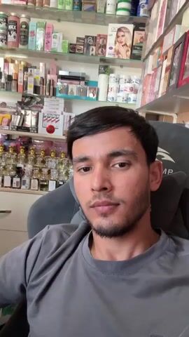 The media features a young man with dark hair and a beard, wearing a grey t-shirt, seated in a grey gaming chair. He is looking directly at the camera and waving. Behind him, shelves are filled with a variety of cosmetic products, including perfumes, lotions, and makeup. Some of the bottles appear to be small perfume samples.

The shelves are organized and well-stocked, suggesting this is a retail environment, likely a shop selling beauty products. The lighting suggests it's daytime, and the indoor setting indicates the weather is not a factor. The man's expression is neutral to slightly smiling, and the wave suggests he is acknowledging the viewer or recording a message.

Visible text includes brand names on the products, such as "WAX," "FASHION," and some in what appears to be Arabic script. The setting is likely a small retail store in Yovon, Tajikistan, given the provided location context, specializing in cosmetics and perfumes. There are no other people visible in the media.