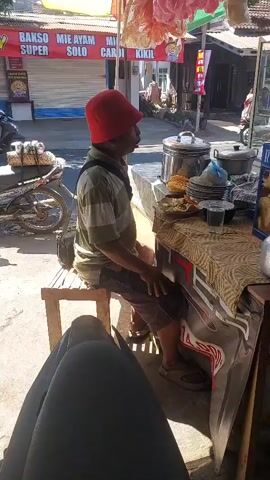 A man wearing a red hat and a striped shirt is sitting on a stool at a food stall. He is looking to his right and extending his left arm forward, as if gesturing or offering something. Behind him, there is a motorcycle parked. The food stall has various items displayed, including what appear to be bowls of food and packaged goods. A woman's legs are visible in the foreground, indicating the perspective of the camera. The scene appears to be outdoors, with sunlight casting shadows.