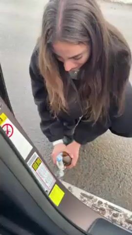 A young woman opens a bottle of champagne against a car door frame. She is wearing a black jacket, a white top, and black pants. The surrounding environment includes snow-covered ground and a road, suggesting a cold or mountainous location. The champagne bottle is brown, and the liquid is effervescent. When opened, champagne spills out, and the bottle cap flies into the car. The woman is smiling and seems excited.