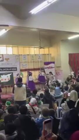 Several young girls are dancing on a stage in a large room. They are wearing matching outfits consisting of white tops, purple skirts, and headbands.  A crowd of people, mostly children, are seated in front of the stage, watching the performance.  Many audience members are holding up their phones to record the event.  Behind the stage is a backdrop with Arabic writing and pictures of Syria. The room is brightly lit with fluorescent lights and has a low ceiling with a ceiling fan. Men in white vests appear to be monitoring the children on stage.