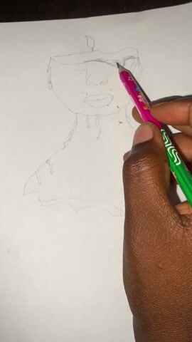 A close-up shot shows a person's dark brown hand holding a pink and green pencil. The hand is sketching the eyebrow of a person with an oval-shaped head, eyes, nose, mouth, and neck. The person being drawn wears what appears to be a hat or some kind of head covering that has a small flame drawn on top. The drawing is on a piece of white paper. The table or surface under the paper appears to be dark brown.