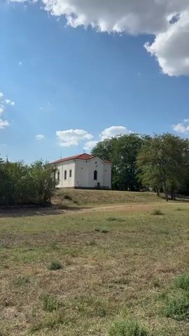 The video shows a small, white, rectangular building with a red tiled roof, which appears to be a church. The building is situated on a grassy hill under a bright blue sky with scattered white clouds. There are trees behind and to the right of the building, and some bushes and dry grass in the foreground. The church has arched windows and crosses on its exterior walls and roof. The camera pans slightly to the right and then zooms in on the church before shifting to a close-up of the sky with sun rays breaking through clouds. No people are visible in the video. The overall scene depicts a peaceful, rural setting on a sunny day.