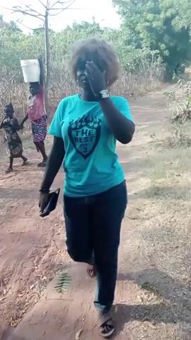 A woman with an afro hairstyle, wearing a blue T-shirt, dark pants, and sandals, is walking on a dirt path in an outdoor setting. She is holding a dark-colored object in her left hand and has a watch on her left wrist. She turns her head to the right and speaks.  In the background, two people walk along the path carrying objects on their heads. Trees and bushes line the sides of the path. The scene is brightly lit.