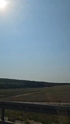 A view of a rural landscape on a sunny day. The sky is clear and blue with the sun shining brightly in the upper left corner. In the middle ground, there are rolling hills and fields, with a line of trees forming a dark green band across the horizon. A guardrail is visible in the foreground, suggesting the footage might be taken from a moving vehicle. The overall impression is one of a peaceful, open natural setting.