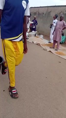 An outdoor street scene unfolds on an unpaved surface next to a plain wall. Approximately ten individuals, primarily adults and one child, are visible. A person wearing a yellow hijab and green dress walks right, while a boy in a light brown outfit walks left. A man in a dark grey top sits on a silver and grey motorcycle parked on the left. Several piles of light-colored grains are spread on two tan tarps on the ground. A man in a white robe holds a camera and smiles. Another man in a light pink robe stands near the grains. A red motorcycle is parked further back against the wall. A black bucket with a yellow object is partially visible in the bottom right foreground.