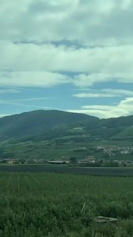 The video shows a landscape view from a moving vehicle. In the foreground, there is a green field with rows of young trees or plants. In the middle ground, there is a town or village with buildings nestled at the base of a large, green mountain. The mountain is covered in vegetation and has terraced slopes, possibly for agriculture. The sky above is partly cloudy with patches of blue visible. The overall impression is of a rural, mountainous region. The video appears to be filmed during the daytime, likely in the late morning or afternoon, given the lighting conditions.