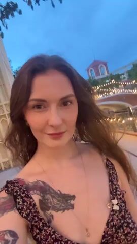 A woman with brown hair and tattoos on her left arm and chest smiles into the camera. She is wearing a dark floral print dress and a gold necklace. She is outdoors at dusk, with a building and string lights visible in the background.