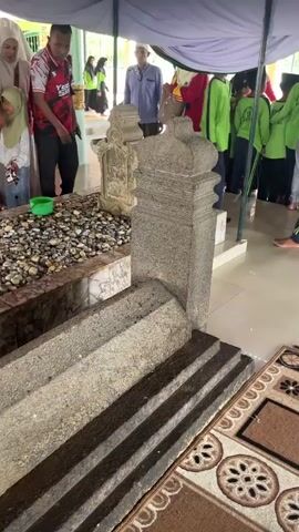 A group of people are gathered around a grave. The grave is made of stone and is covered with pebbles. There are several people standing around the grave, some of whom are holding green buckets. The people are dressed in a variety of clothing, including hijabs and t-shirts. The environment appears to be outdoors, with a canopy providing shade. The camera is focused on the grave, with the people in the background.
