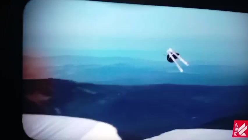 A skier in mid-air, performing a jump against a backdrop of blue mountains. The skier is wearing black and white. The second half of the video shows a different scene with a person on skis falling onto a snowy slope near a red flag. There is also a graphic in the corner that says "THE ULTIMATE RACE" and "SUNDAY - JULY 30, 20:30 CEST".