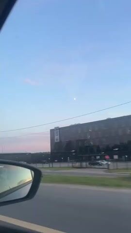 This media file captures a view from inside a moving vehicle, likely a car, as it travels along a road in Korobovo, Russia. The scene is set during a beautiful twilight, with a clear blue sky transitioning into soft hues of pink and orange near the horizon, indicating either sunrise or sunset. A faint, small moon is visible high in the sky.

The primary subject is a large, modern building with a dark facade, situated across the road. The building has a prominent sign displaying Cyrillic text that appears to read "ШИНОМОНТАЖ МИР ШИН И ДИСКОВ". This translates to "Tire Fitting World of Tires and Wheels," suggesting it is a tire service center or auto shop. The building's windows are illuminated, and displays of tires and wheels can be seen inside.

To the left of this main building, another, longer industrial-style building is visible, also appearing dark. A street light stands near the edge of the road, and power lines crisscross the sky. In the reflection of the car's side mirror, a white car and the road ahead can be seen, further confirming the vehicle's movement. The overall mood of the media is serene and calm, enhanced by the soft, diffused light of twilight and the peaceful, expansive sky. The environment appears to be an urban or suburban setting, characterized by commercial buildings and roadways.