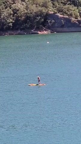 A person is paddleboarding on a large body of water. The person is standing on a paddleboard and is holding a paddle. The water is blue and calm. In the background, there is a shoreline with trees and a hill. There is a white spherical object floating in the water in the distance. The sun is shining and the sky is clear. The video is shot from a distance, and the details of the person and the paddleboard are not clear.
