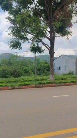 The video shows a roadside view of a rural area. The road is paved with yellow dividing lines. There are low buildings on the side of the road, including one with a red roof and another painted a light color. Several motorbikes are parked near one of the buildings. Trees and green vegetation line the road, and in the background, there are visible mountains against a sky with scattered clouds. The camera appears to be moving along the road.