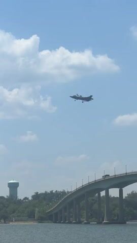 The video shows a fighter jet flying in the sky, presumably on approach to landing. The plane is gray and the landing gear is deployed. In the background are trees, a body of water, a bridge, a water tower, light poles, and the top of a boat. The sky is blue with some white clouds.